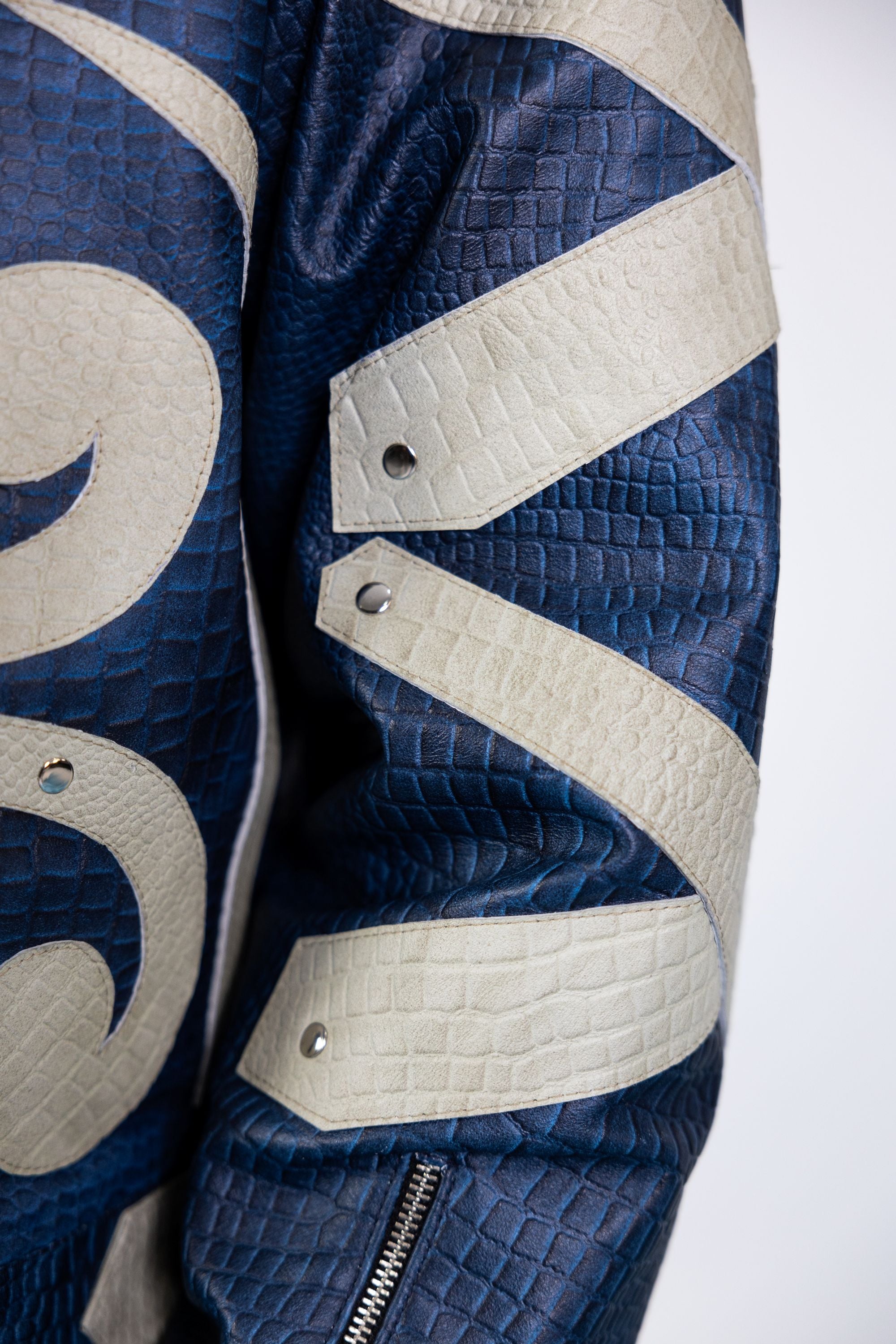 Close-up of blue embossed leather jacket highlighting abstract beige appliqué swirls and metal rivet detailing