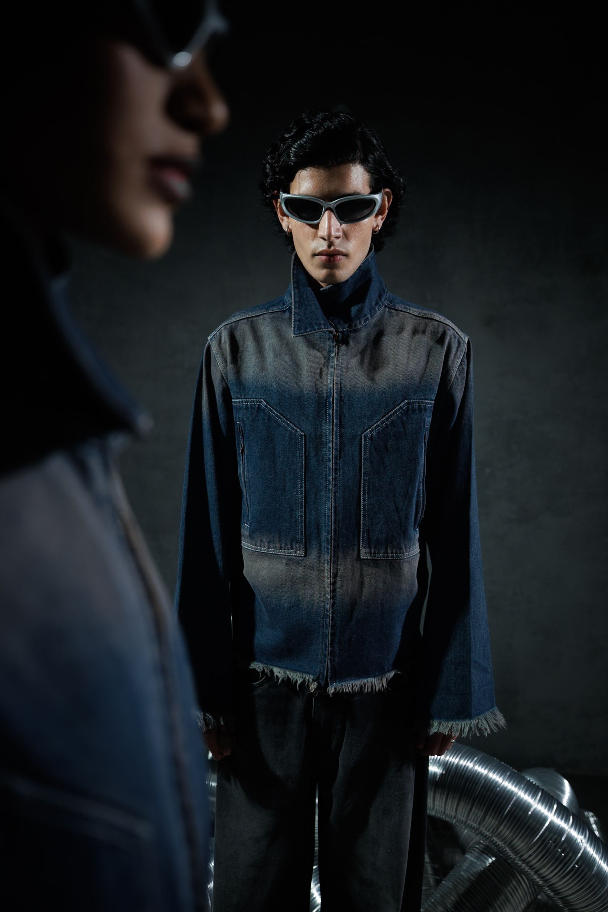 Model wearing a blue denim jacket with zipper front and sunglasses, styled against a dark background